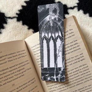 May include: Black and white bookmark featuring a gothic arched window design with floral accents. The bookmark is rectangular and placed on an open book. The image is in a high-angle shot.