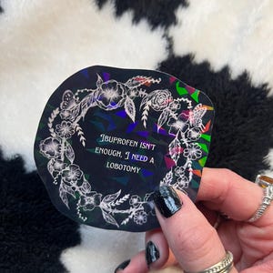 May include: A holographic sticker with a floral wreath design. The sticker has the text "Ibuprofen isn't enough, I need a lobotomy" in white. The sticker is held up by a person with black nail polish.