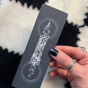 May include: A black bookmark featuring a white illustration of a candle with two radiating suns. The candle is in the center, with a flame at the top and a smaller flame at the bottom. The bookmark is rectangular and held by a hand with black nail polish.
