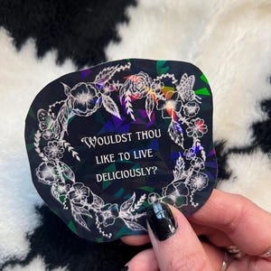 May include: A holographic sticker with a dark background and a floral wreath design. The sticker features the text "WOULDST THOU LIKE TO LIVE DELICIOUSLY?" in white. The sticker has a unique, iridescent finish.