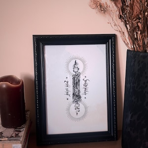 May include: Black framed art print featuring a detailed illustration of a candle with radiating lines and stylized text. The artwork is set against a white background and is displayed on a wooden surface. A dark brown candle and books are in the foreground.