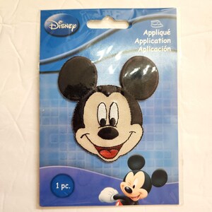 May include: A Disney Mickey Mouse embroidered iron-on patch in a retail package. The patch features the character's face with black ears, a cream-colored face, and a red mouth. The blue packaging includes the text Disney, Appliqué, Application, and 1 pc.