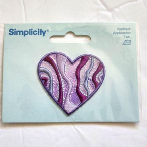 May include: A Simplicity brand heart-shaped embroidered appliqué patch featuring a wavy, abstract pattern in shades of purple, lavender, and silver. The patch is mounted on a light blue cardboard backing with the text Simplicity and Appliqué 1 pc.