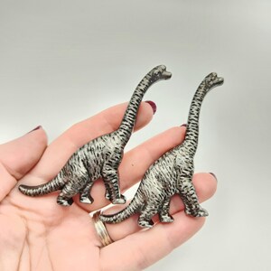 May include: Two silver dinosaur figurines with long necks and striped patterns. The figurines are Brachiosaurus-like in design, with four legs and a long tail. The dinosaurs are held in a hand against a white background.