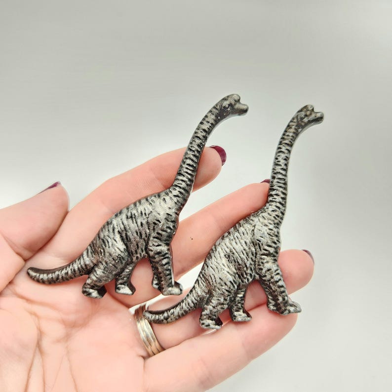 May include: Two silver dinosaur figurines with long necks and striped patterns. The figurines are Brachiosaurus-like in design, with four legs and a long tail. The dinosaurs are held in a hand against a white background.