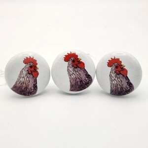 May include: Three white cabinet knobs with a rooster design. Each knob features a detailed illustration of a rooster's head with a red comb and wattle, set against a white background. The rooster's feathers are depicted in shades of gray and brown.