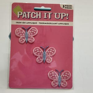 May include: A package of three iron-on butterfly appliqués. Each butterfly has pink wings with white polka dots and a light blue body. The package is pink with the text "PATCH IT UP!" and "3 PIECES".