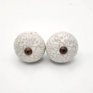 May include: Two white ceramic cabinet knobs with a floral and swirl design. Each knob has a dark brown center and a decorative pattern in a lighter shade of white. These knobs are suitable for furniture or cabinet upgrades.