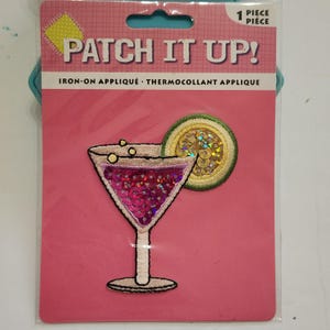 May include: Iron-on applique patch featuring a cocktail design. The patch has a pink background with the words "PATCH IT UP!" and "IRON-ON APPLIQUE". The cocktail glass is filled with pink sequins and has a lime slice.