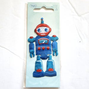 May include: An embroidered robot applique patch by Wrights on a light blue cardboard backing. The robot features a red head, blue body, and blue limbs with red accents, including a chest panel with a white gauge and three red circular buttons.