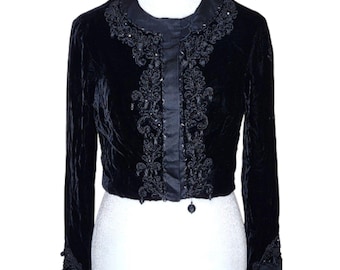 May include: A black velvet cropped jacket featuring long sleeves and a round neckline. The front placket and collar area are adorned with intricate black embroidery and beadwork, with additional bead detailing on the sleeve cuffs.