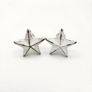 May include: Two white, star-shaped cabinet knobs with a distressed finish. The knobs have a five-point star design and are made of metal. The knobs are ideal for adding a rustic touch to furniture or decor.