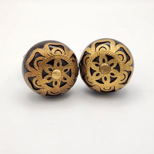 May include: Two decorative cabinet knobs. Each knob is a dark brown sphere with a gold-colored, openwork floral design overlay. The center of each knob features a small, gold-colored circular accent. The knobs are likely made of wood and metal.