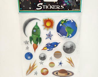 May include: A clear plastic package containing 21 space-themed stickers. The header reads "Nicole Quality Value" and "STICKERS". The stickers feature a green rocket, a blue rocket, the Earth, Saturn, a crescent moon, stars, and various planets.