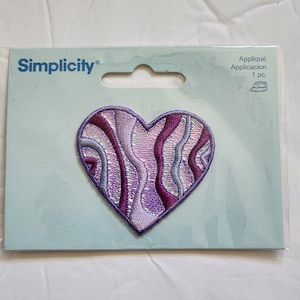 May include: A heart-shaped applique patch with a swirling design in shades of purple, lavender, and silver. The patch is on a light blue backing with the brand name "Simplicity" printed in blue. The package also includes the text "Appliqué Application 1 pc."