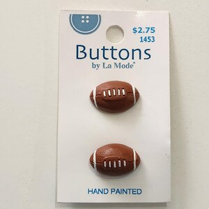 May include: Two brown football-shaped buttons with white stripes and stitching are displayed on a white card. The card reads "Buttons by La Mode" and "Hand Painted". The buttons are designed to resemble miniature footballs.