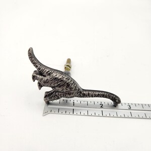 May include: A silver-coloured dinosaur-shaped cabinet knob with a bolt. The knob is approximately 7.6 cm long. The dinosaur has a textured, striped pattern. The knob is on a white background.