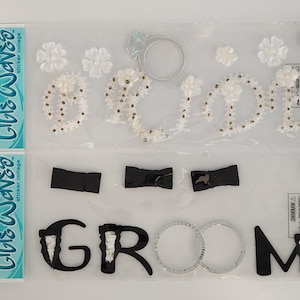 May include: Clear plastic package containing wedding-themed stickers. The stickers include the word "Bride" in white beaded letters, a ring, white flowers, and the word "Groom" in black letters with silver rings and black bow tie accents.