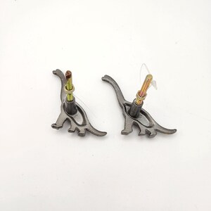 May include: Two grey metal dinosaur-shaped cabinet knobs with attached bolts. The knobs feature a stylised dinosaur design and are set against a white background. The bolts are gold and silver in colour.