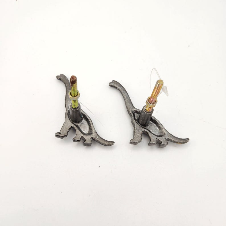 May include: Two grey metal dinosaur-shaped cabinet knobs with attached bolts. The knobs feature a stylised dinosaur design and are set against a white background. The bolts are gold and silver in colour.