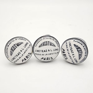 May include: Three white and black ceramic cabinet knobs with a circular design. Each knob features text in French, including "J. Metraz & L. Long, Paris". The knobs have a vintage, antique aesthetic.