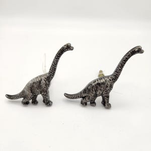 May include: Two silver-colored Brachiosaurus dinosaur figurines. The dinosaurs have long necks and tails, with textured detailing on their bodies. They are standing on a white surface.