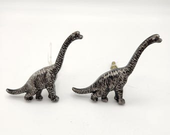 May include: Two silver-colored Brachiosaurus dinosaur figurines. The dinosaurs have long necks and tails, with textured detailing on their bodies. They are standing on a white surface.