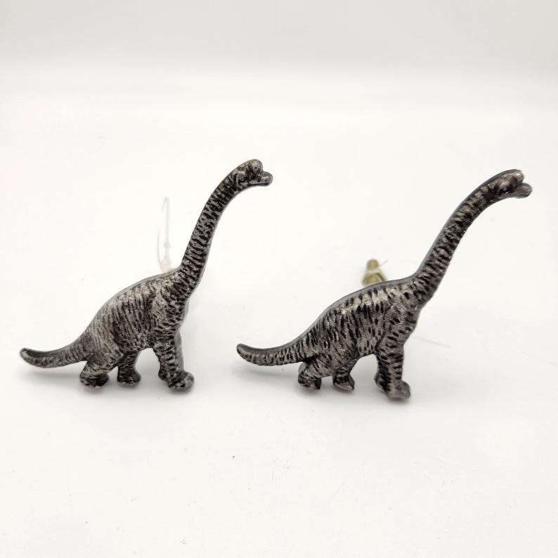 May include: Two silver-coloured Brachiosaurus dinosaur figurines. The dinosaurs have long necks and tails, with textured detailing on their bodies. They are standing on a white surface.