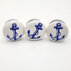 May include: Three white ceramic cabinet knobs with a navy blue anchor design. Each knob has a silver-colored metal base. The anchor is centered on the knob and is a classic nautical symbol.