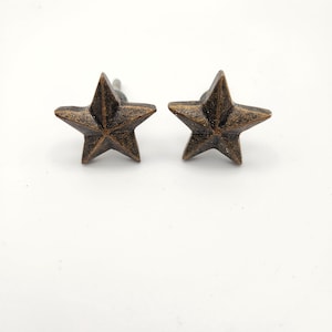 May include: Two bronze-colored star-shaped cabinet knobs. Each knob has a five-pointed star design with a distressed finish. The knobs are mounted on silver-colored stems, suitable for furniture or decorative projects.