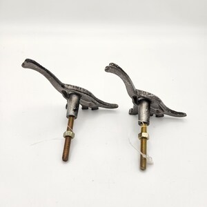 May include: Two cast iron dinosaur-shaped cabinet pulls. The pulls are dark grey with a textured finish. Each pull has a long neck and tail, with a threaded rod and nut for mounting. These decorative pulls are ideal for a child's room or a dinosaur-themed space.