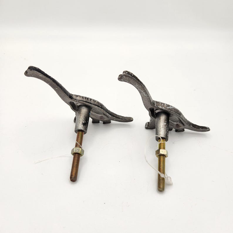 May include: Two cast iron dinosaur-shaped cabinet pulls. The pulls are dark grey with a textured finish. Each pull has a long neck and tail, with a threaded rod and nut for mounting. These decorative pulls are ideal for a child's room or a dinosaur-themed space.