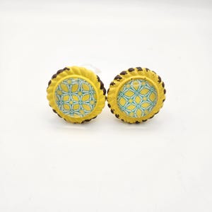 May include: Two yellow cabinet knobs with a distressed finish. Each knob features a decorative, patterned center in yellow and teal, surrounded by a yellow border and a dark brown rope-like edge. The knobs are designed for furniture.