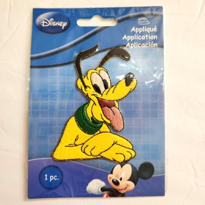 May include: A Disney Pluto iron-on embroidered patch featuring the yellow dog with a green collar and pink tongue, packaged on a blue card with the text Appliqué Application Aplicación and 1 pc. A small Mickey Mouse graphic is at the bottom.