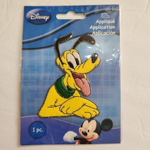 May include: A Disney Pluto applique patch on a blue background. The patch features a yellow Pluto with a green collar and a pink tongue sticking out. The packaging includes the Disney logo and the text "1 pc."