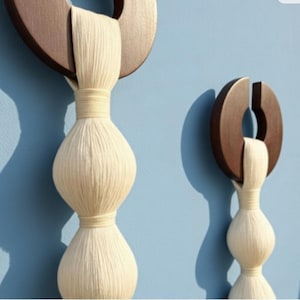 May include: Two decorative wall hangings. Each features a dark brown wooden ring with a section cut out, and a cream-colored, vertically-oriented, double-bulb shape made of tightly wound yarn. The background is a light blue.