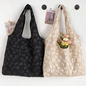Pu&ograve; includere: Due borse tote, una nera e una beige, ciascuna decorata con un motivo floreale. La borsa beige contiene un mazzo di fiori. Entrambe le borse sono appese a ganci neri su un muro bianco.