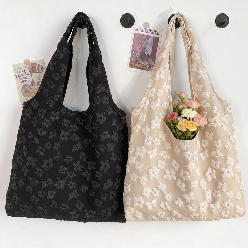 Pu&ograve; includere: Due borse tote, una nera e una beige, ciascuna decorata con un motivo floreale. La borsa beige contiene un mazzo di fiori. Entrambe le borse sono appese a ganci neri su un muro bianco.