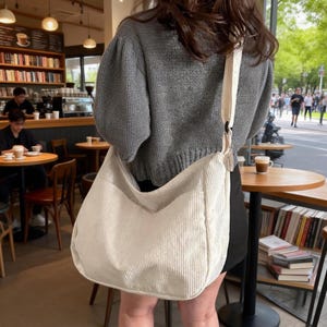 Puede incluir: Un bolso de hombro de pana color crema con una correa ancha. El bolso se lleva sobre el hombro. El fondo muestra un ambiente de cafeter&iacute;a con mesas, sillas y estanter&iacute;as.