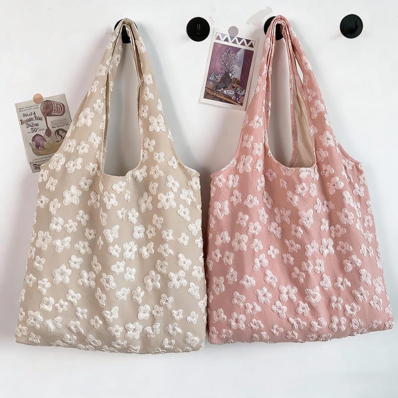 Pu&ograve; includere: Due borse tote floreali, una beige e una rosa, sono appese a una parete bianca. Le borse presentano motivi floreali bianchi. Sono appese a ganci neri. Una piccola immagine e un foglio di carta sono anche attaccati al muro.