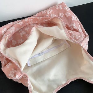 Pu&ograve; includere: Una borsa tote rosa con un motivo floreale. La borsa ha un esterno rosa chiaro con ricami bianchi a forma di fiore. L'interno &egrave; color crema chiaro con una tasca con cerniera. La borsa &egrave; aperta, rivelando l'interno.