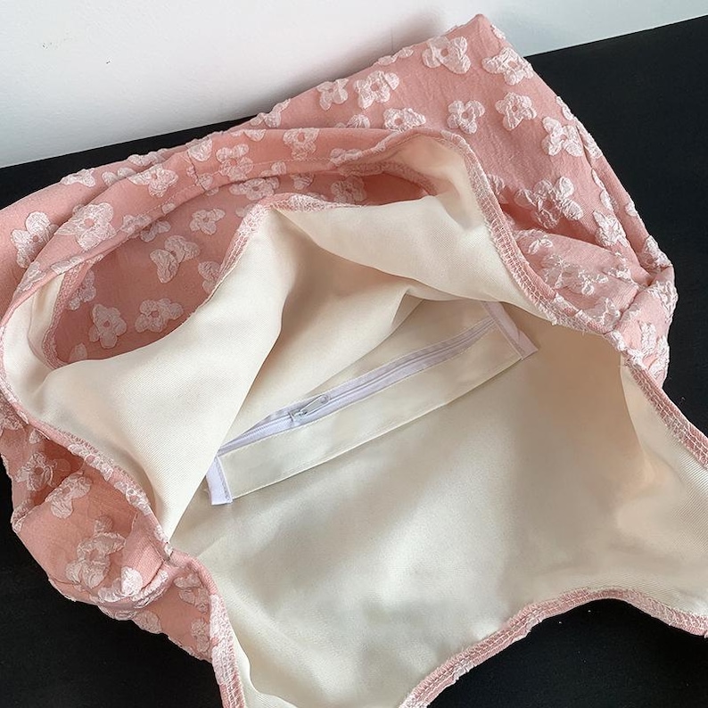 Pu&ograve; includere: Una borsa tote rosa con un motivo floreale. La borsa ha un esterno rosa chiaro con ricami bianchi a forma di fiore. L'interno &egrave; color crema chiaro con una tasca con cerniera. La borsa &egrave; aperta, rivelando l'interno.