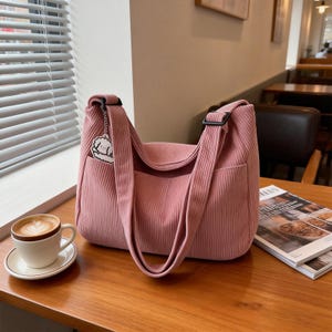 Puede incluir: Bolso de hombro de pana rosa empolvado con un compartimento principal grande y un bolsillo exterior. El bolso tiene una correa ancha y un peque&ntilde;o dije. Una taza de caf&eacute; est&aacute; sobre una mesa.