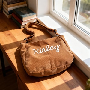 Puede incluir: Bolso de hombro de pana marr&oacute;n con el nombre "Kinley" bordado en escritura blanca. El bolso tiene una correa larga y un bolsillo delantero. El bolso est&aacute; sobre una superficie de madera cerca de una ventana.