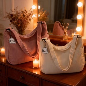 Puede incluir: Dos bolsos de hombro de pana, uno rosa y otro crema, sobre una superficie de madera. Cada bolso tiene una correa larga y un peque&ntilde;o adorno. Se ven velas encendidas y un jarr&oacute;n con flores secas.