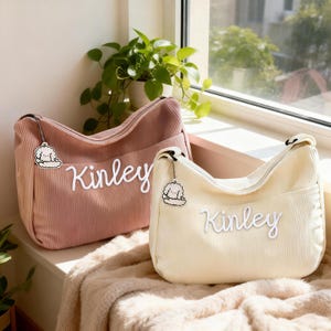 Puede incluir: Dos bolsos de hombro de pana, uno rosa empolvado y otro crema, se muestran uno al lado del otro. Cada bolso presenta el nombre "Kinley" en escritura blanca y un charm con un perro de dibujos animados. Los bolsos tienen cremallera y un bolsillo delantero.