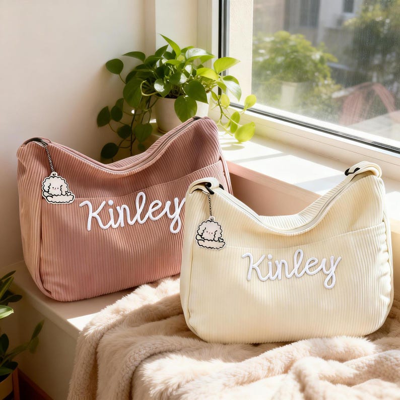 Puede incluir: Dos bolsos de hombro de pana, uno rosa empolvado y otro crema, se muestran uno al lado del otro. Cada bolso presenta el nombre "Kinley" en escritura blanca y un charm con un perro de dibujos animados. Los bolsos tienen cremallera y un bolsillo delantero.