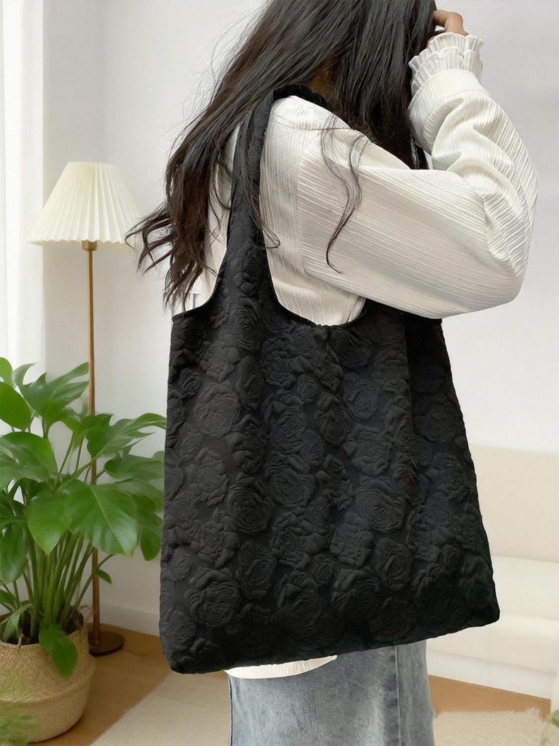 Pu&ograve; includere: Borsa tote nera con motivo a rose in rilievo. La borsa &egrave; realizzata in tessuto strutturato e ha una grande capacit&agrave;. Viene portata a spalla. Lo sfondo comprende una camicia bianca e una pianta.