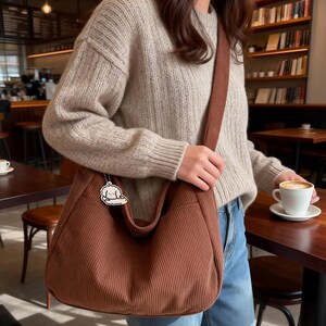 Puede incluir: Bolso de hombro de pana marr&oacute;n con una correa a juego. El bolso tiene un compartimento principal grande y un peque&ntilde;o colgante. La persona lleva un jersey beige y vaqueros azules. Una taza de caf&eacute; est&aacute; sobre una mesa de madera.
