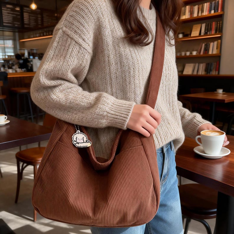 Puede incluir: Bolso de hombro de pana marr&oacute;n con una correa a juego. El bolso tiene un compartimento principal grande y un peque&ntilde;o colgante. La persona lleva un jersey beige y vaqueros azules. Una taza de caf&eacute; est&aacute; sobre una mesa de madera.
